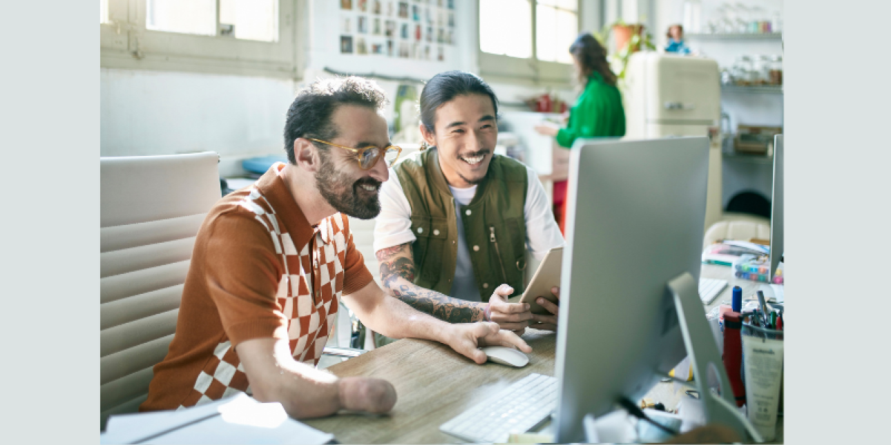 A white man with dark hair and beard and an Asian man with a dark ponytail, are sat together laughing at a computer screen in a creative studio.